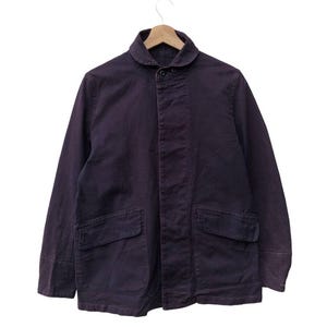 May include: A dark purple long-sleeve jacket featuring a rounded collar, a button-up front closure with a concealed placket, and two large rectangular patch pockets on the lower front. The garment is displayed on a wooden hanger.