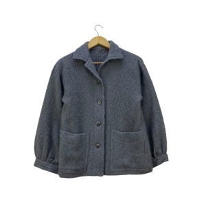 May include: A gray wool jacket with a collared neckline, three buttons, and two front pockets. The jacket is hanging on a wooden hanger against a white background. The sleeves are slightly puffed.