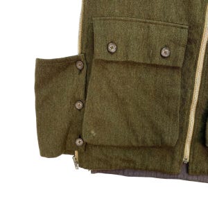 May include: Close-up of a green wool vest with multiple pockets and button closures. The vest has a zipper closure and a brown lining. The pockets are rectangular and have button closures. The vest is made of a thick, textured wool fabric.