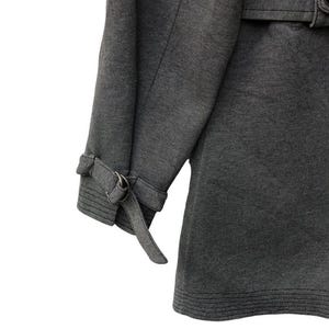 May include: Close-up of a gray coat sleeve with a buckle detail at the cuff. The coat has a textured fabric and a belt detail at the waist. The coat is a neutral color and appears to be made of a durable material.