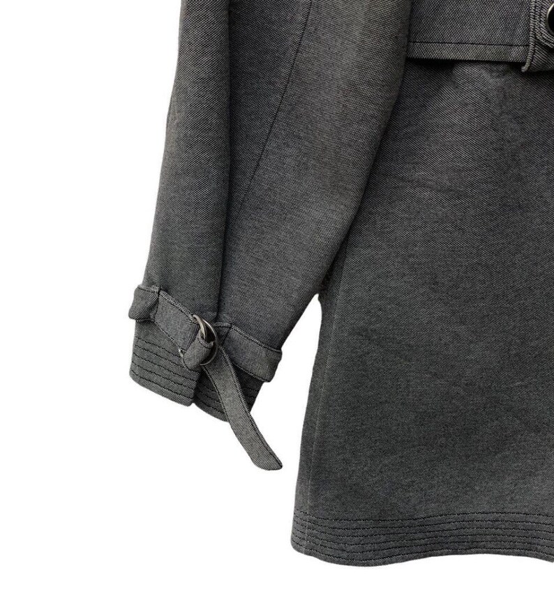May include: Close-up of a gray coat sleeve with a buckle detail at the cuff. The coat has a textured fabric and a belt detail at the waist. The coat is a neutral color and appears to be made of a durable material.