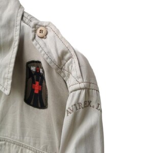 May include: Close-up of a tan military-style shirt. The shirt features a shoulder epaulet with a button, a patch with a red cross and the word "MEDIC", and the embroidered brand name "AVIREX".