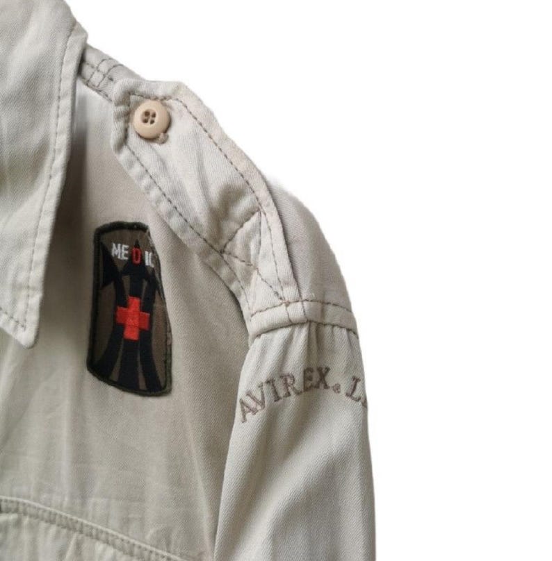 May include: Close-up of a tan military-style shirt. The shirt features a shoulder epaulet with a button, a patch with a red cross and the word "MEDIC", and the embroidered brand name "AVIREX".