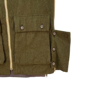 May include: Close-up of a green wool vest with multiple pockets and button closures. The vest has a full-length zipper and a brown lining. The pockets are rectangular and have button closures. The vest is made of a thick, textured wool fabric.