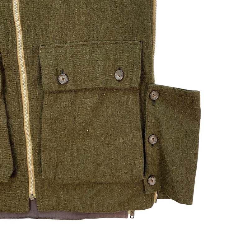 May include: Close-up of a green wool vest with multiple pockets and button closures. The vest has a full-length zipper and a brown lining. The pockets are rectangular and have button closures. The vest is made of a thick, textured wool fabric.