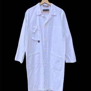Puede incluir: Una bata de laboratorio larga y blanca con cuello, cierre de botones y múltiples bolsillos. La bata está colgada en una percha de madera sobre un fondo negro. La bata tiene un pequeño logotipo en el bolsillo izquierdo.