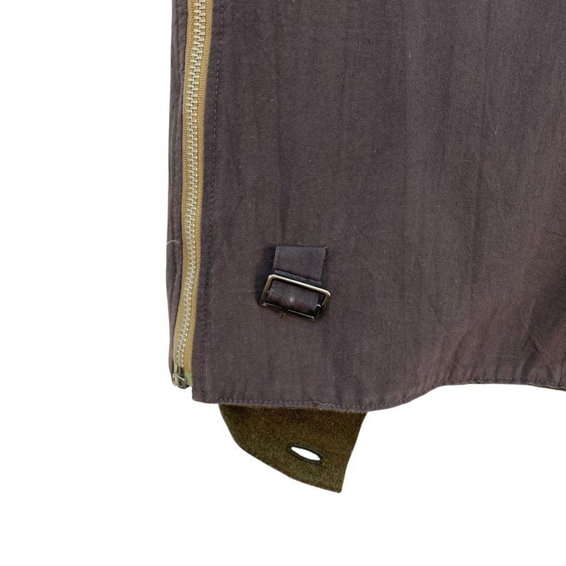 May include: Close-up of a brown garment with a tan zipper and a small leather strap with a metal buckle. The bottom edge has a green fabric detail with a buttonhole.