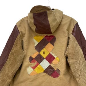 May include: Tan jacket with brown leather detailing and a patchwork teddy bear design on the back. The jacket has a hood and a stitched bear graphic with a letter "K" in the centre. The sleeves are brown leather.