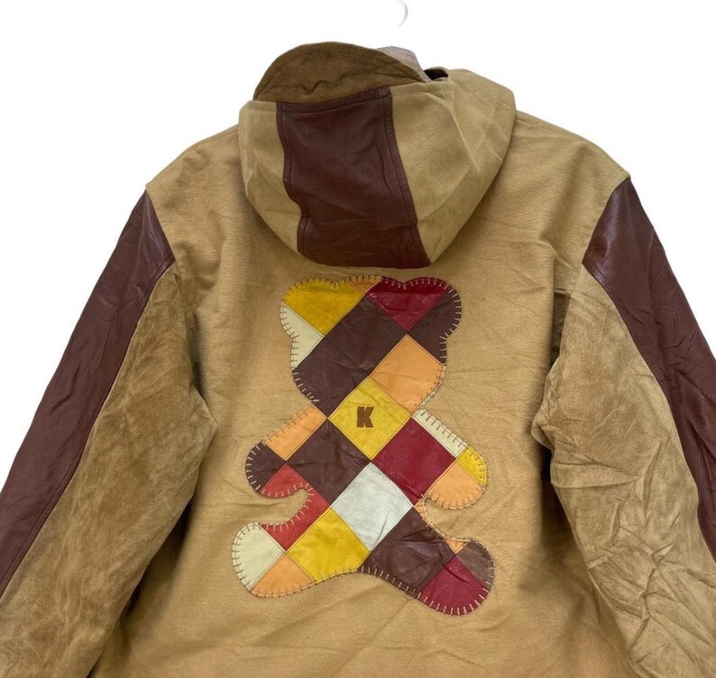 May include: Tan jacket with brown leather detailing and a patchwork teddy bear design on the back. The jacket has a hood and a stitched bear graphic with a letter "K" in the centre. The sleeves are brown leather.