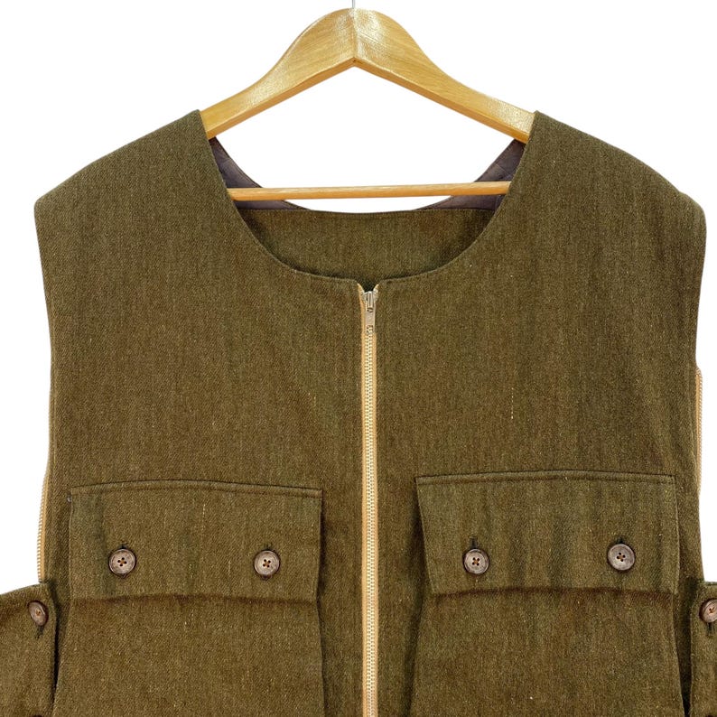 May include: Olive green vest with a full-length zipper and two front pockets with wooden button closures. The vest is sleeveless and hangs on a wooden hanger. The fabric has a textured appearance.