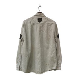 May include: A light beige button-down shirt with long sleeves, hanging on a black hanger. The shirt features dark patches on the shoulders and sleeves. The back of the shirt is visible, showing the collar and stitching details.