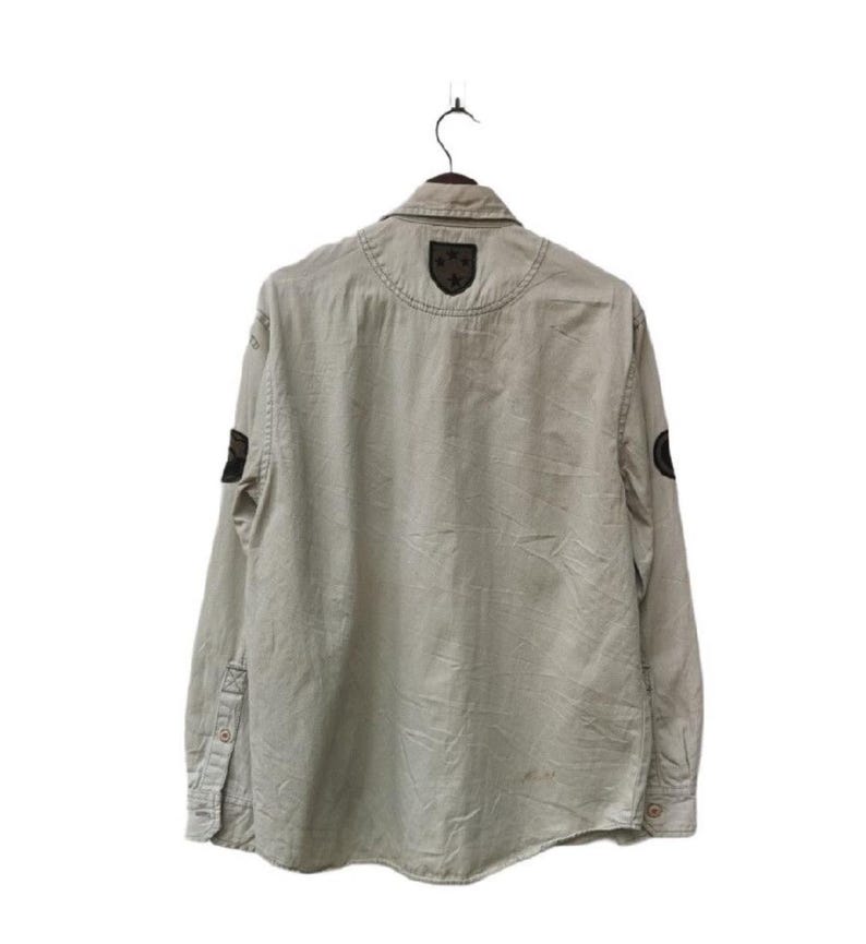 May include: A light beige button-down shirt with long sleeves, hanging on a black hanger. The shirt features dark patches on the shoulders and sleeves. The back of the shirt is visible, showing the collar and stitching details.