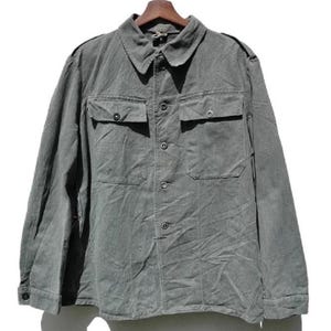 Puede incluir: Una camisa gris abotonada con dos bolsillos en el pecho y un cuello, colgada de una percha de madera. La camisa de manga larga tiene cierres de botones en la parte delantera y en los puños. La tela parece ser un material duradero, posiblemente de estilo militar.