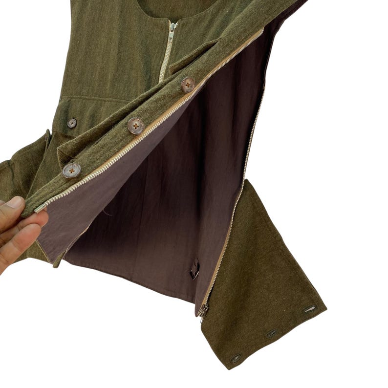 May include: Olive green vest with a brown interior. The vest features a zipper closure, wooden buttons, and small pockets. The interior lining is a solid brown color. The vest is made of a textured fabric.