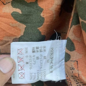May include: Close-up of a camouflage pattern fabric in shades of orange, brown, and green. A white tag with Japanese text and care symbols is attached. The tag includes the text "P0303FHC16 B".