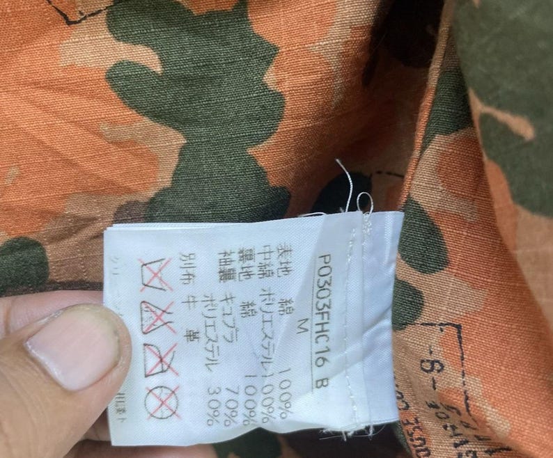 May include: Close-up of a camouflage pattern fabric in shades of orange, brown, and green. A white tag with Japanese text and care symbols is attached. The tag includes the text "P0303FHC16 B".
