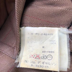 Może przedstawiać: Zbliżenie brązowej tkaniny z cienkimi czerwonymi paskami. Dołączona biała metka z tekstem, w tym "PC-04010M", "M", "AD1996" i japońskimi znakami. Etykieta zawiera r&oacute;wnież symbole pielęgnacyjne.
