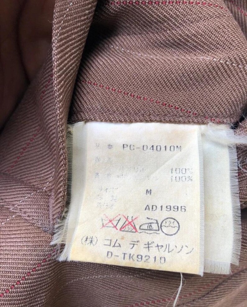 Może przedstawiać: Zbliżenie brązowej tkaniny z cienkimi czerwonymi paskami. Dołączona biała metka z tekstem, w tym "PC-04010M", "M", "AD1996" i japońskimi znakami. Etykieta zawiera r&oacute;wnież symbole pielęgnacyjne.