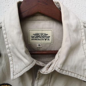 May include: Close-up of a tan jacket with a collared neckline and button closure. The jacket features a label with text including "AVIREX LTD" and "EQUIPMENT-TYPE-AVIREX". The jacket is hanging on a wooden hanger.