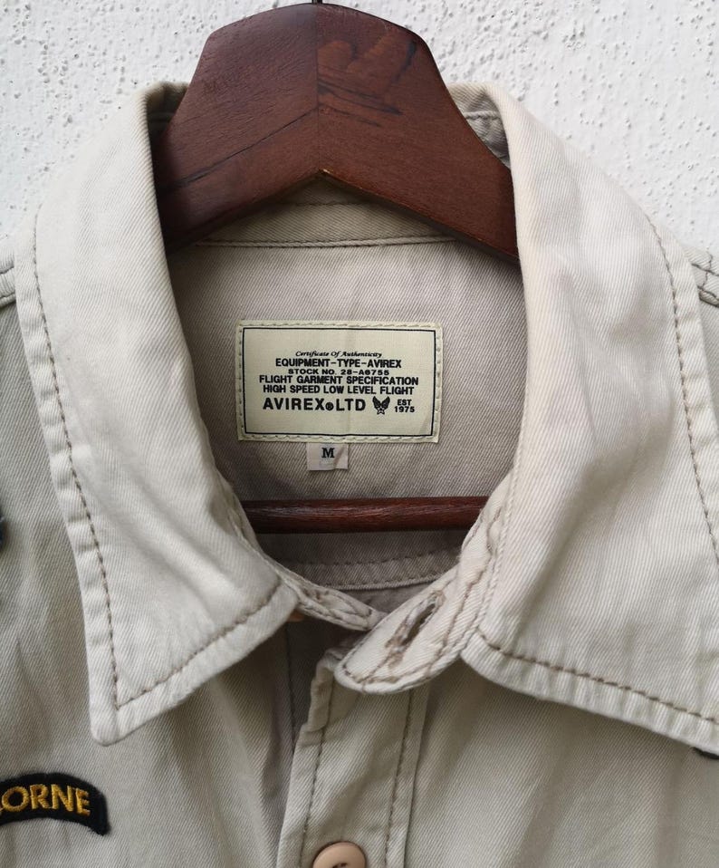 May include: Close-up of a tan jacket with a collared neckline and button closure. The jacket features a label with text including "AVIREX LTD" and "EQUIPMENT-TYPE-AVIREX". The jacket is hanging on a wooden hanger.