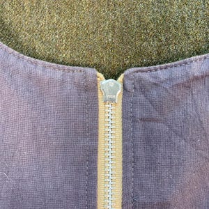 May include: Close-up of a garment with a visible zipper. The zipper is gold-colored and has the brand name "YKK" stamped on the pull tab. The fabric is a muted purple color, with a contrasting tan-colored strip where the zipper is sewn.