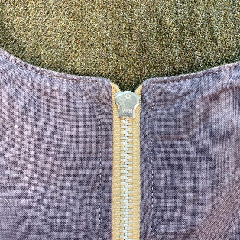 May include: Close-up of a garment with a visible zipper. The zipper is gold-colored and has the brand name "YKK" stamped on the pull tab. The fabric is a muted purple color, with a contrasting tan-colored strip where the zipper is sewn.