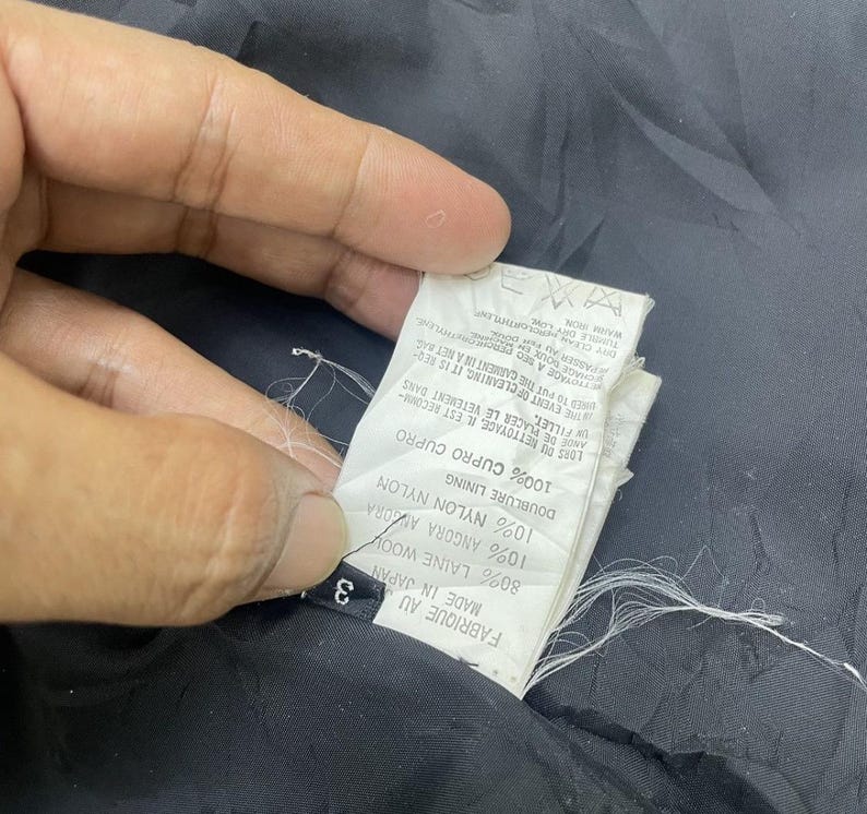 Puede incluir: Una etiqueta de tela negra con texto blanco que detalla la composici&oacute;n de la tela de la prenda. La etiqueta dice "Fabrique au Japan Made in Japan 3 80% Laine Wool 10% Angora Amora Doublure Lining 10% Nylon Nylon 100% Cupro Cupro On Fillet Ande de Placer le Vetement dans The Event of Cleaning, It is Req Need to Put the Carment in Anet Mag Yoyage a Seo Prometruthylene Passeriad F Dry Clean Warm Iron"