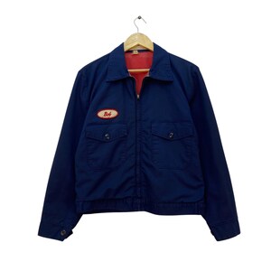 Könnte beinhalten: Marineblaue Arbeitsjacke mit rotem Innenfutter. Die Jacke hat einen Reißverschluss, zwei Brusttaschen und einen kleinen Aufnäher mit der Aufschrift "Bob". Sie hängt an einem Holzbügel.