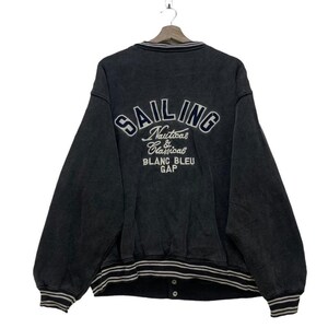 Könnte beinhalten: Ein dunkelgraues Sweatshirt mit dem Wort "SAILING" in Weiß auf dem Rücken gestickt. Darunter steht der Text "Nautical & Classical BLANC BLEU GAP". Das Sweatshirt hat Streifen am Kragen, an den Bündchen und am Saum.