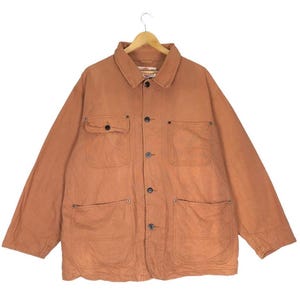 Puede incluir: Chaqueta de trabajo color óxido con cuatro bolsillos delanteros y cierre de botones. La chaqueta tiene cuello y mangas largas. La tela parece ser de algodón o lona duradera, adecuada para actividades al aire libre.