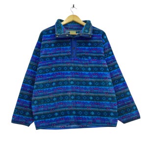 May include: A long-sleeve pullover fleece featuring a vibrant blue, teal, and purple geometric pattern. The garment includes a high collar with a snap-button placket, long sleeves, and a straight hemline, all displayed on a wooden hanger.