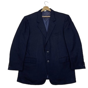 Puede incluir: Un blazer azul marino con cierre clásico de dos botones, solapas con muescas y dos bolsillos delanteros. El blazer está colgado de una percha de madera. Las mangas tienen botones en los puños. El blazer es de color sólido.