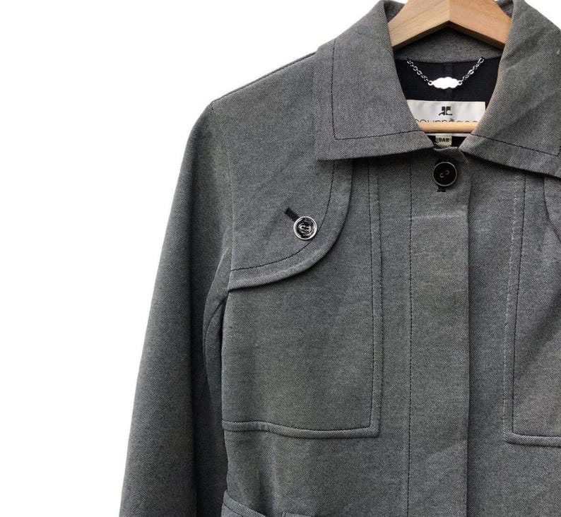 May include: A gray coat with a notched collar and a button closure. The coat has two front pockets and a small decorative button on the left side. The coat is hanging on a wooden hanger.