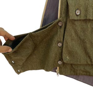 May include: Close-up of a green wool vest with a brown interior lining. The vest features a zipper, button closures, and a pocket. The buttons are brown with a cross design. The vest is made of a textured fabric.