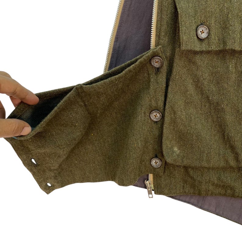 May include: Close-up of a green wool vest with a brown interior lining. The vest features a zipper, button closures, and a pocket. The buttons are brown with a cross design. The vest is made of a textured fabric.