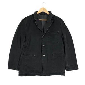 Op de afbeelding: Een zwarte denim blazer met een klassiek design, met een gekerfde revers, drie knopen en twee voorzakken. De jas hangt aan een houten hanger.