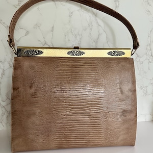 May include: A vintage brown handbag with a textured exterior and a curved handle. The bag features a gold-toned clasp with decorative cutouts. The handle is a darker brown, complementing the bag's overall color scheme.
