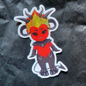May include: A sticker featuring a cartoon devil with red skin, black horns, and a glittery gold and red head. The devil has a red chest and black legs and arms. The sticker has the website dollbite.com printed at the bottom.