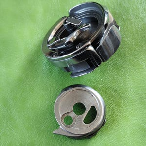 May include: Two silver sewing machine bobbins. One is a round bobbin with a hole in the center. The other is a larger bobbin with a metal hook and a small lever.