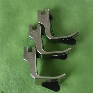 Peut inclure: Trois pieds-de-biche de machine à coudre en argent avec des pieds en plastique noir. Les pieds sont étiquetés avec les numéros 364465, 364465 et 364467.
