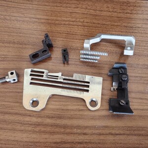Pode incluir: Um conjunto de peças de máquina de costura, incluindo um pé de metal, uma placa de agulha de metal, um calcador de metal e duas peças de plástico preto.