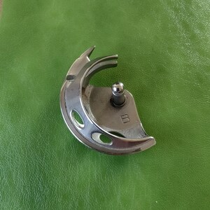Pode incluir: Um pé de máquina de costura de prata com uma abertura circular e uma pequena manivela de metal. O pé é marcado com a letra "B".