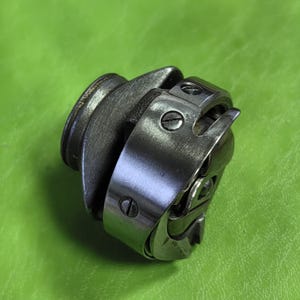 May include: A silver sewing machine bobbin case with a metal hook and a screw on the side.