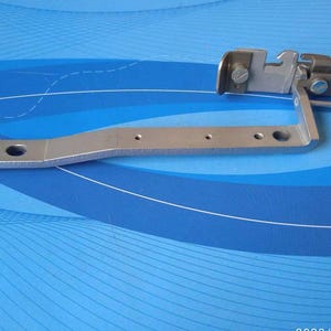 Può includere: Un pezzo di macchina da cucire in metallo argentato con un braccio curvo e un piccolo pezzo rettangolare incernierato con una vite. Il pezzo è su uno sfondo blu.