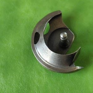 May include: A silver sewing machine bobbin case with a small hole on the side and a metal hook.