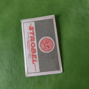 May include: Rectangular box with the word "STROBEL" in large red letters, and a red circular emblem. The box has a gray and white color scheme. The text "Spezial Nähmaschinen Fabrik München" is also visible.