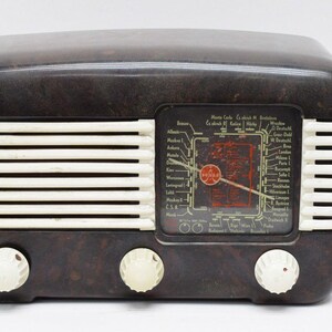 Może przedstawiać: Antyczne radio z bakelitu w kolorze brązowym z białą kratką głośnika i tarczą z nazwami stacji i częstotliwościami. Radio ma trzy pokrętła z przodu.