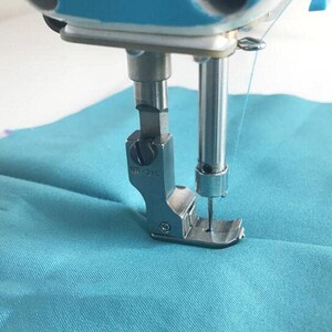 Pode incluir: Close-up de um pé de máquina de costura com uma agulha e linha passando por um tecido turquesa. O pé está etiquetado como "NR-215".