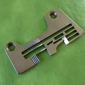 Op de afbeelding: Een zilveren metalen naaimachinevoet met een reeks groeven en een klein gaatje bovenaan.