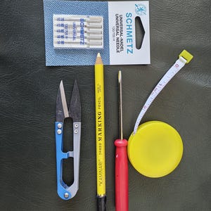 Peut inclure: Un ensemble d'outils de couture comprenant une paire de ciseaux à manche bleu, un crayon jaune avec l'inscription "KAMINARI" DRESS MARKING PENCIL", un tournevis rouge, un mètre ruban jaune et un paquet d'aiguilles étiqueté "SCHMETZ UNIVERSAL-NADEL UNIVERSAL NEEDLE 130/705 H".